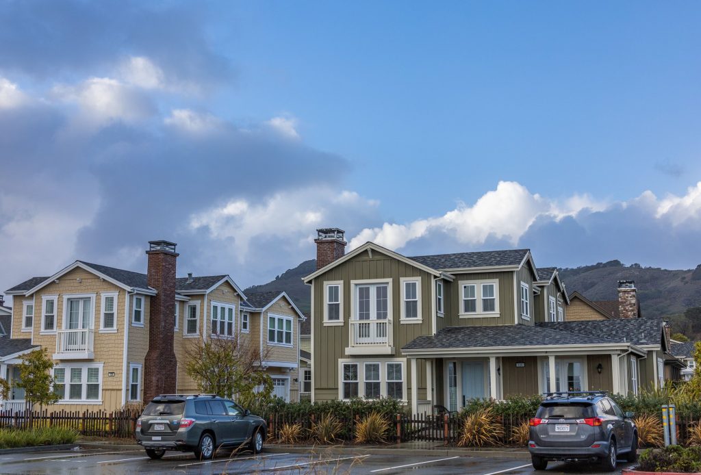 A street of two-story suburban houses under a cloudy sky with cars parked along the kerb