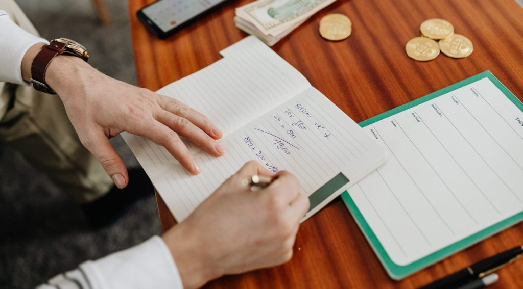 Hands working through figures in a notebook with banknotes and coins on the table